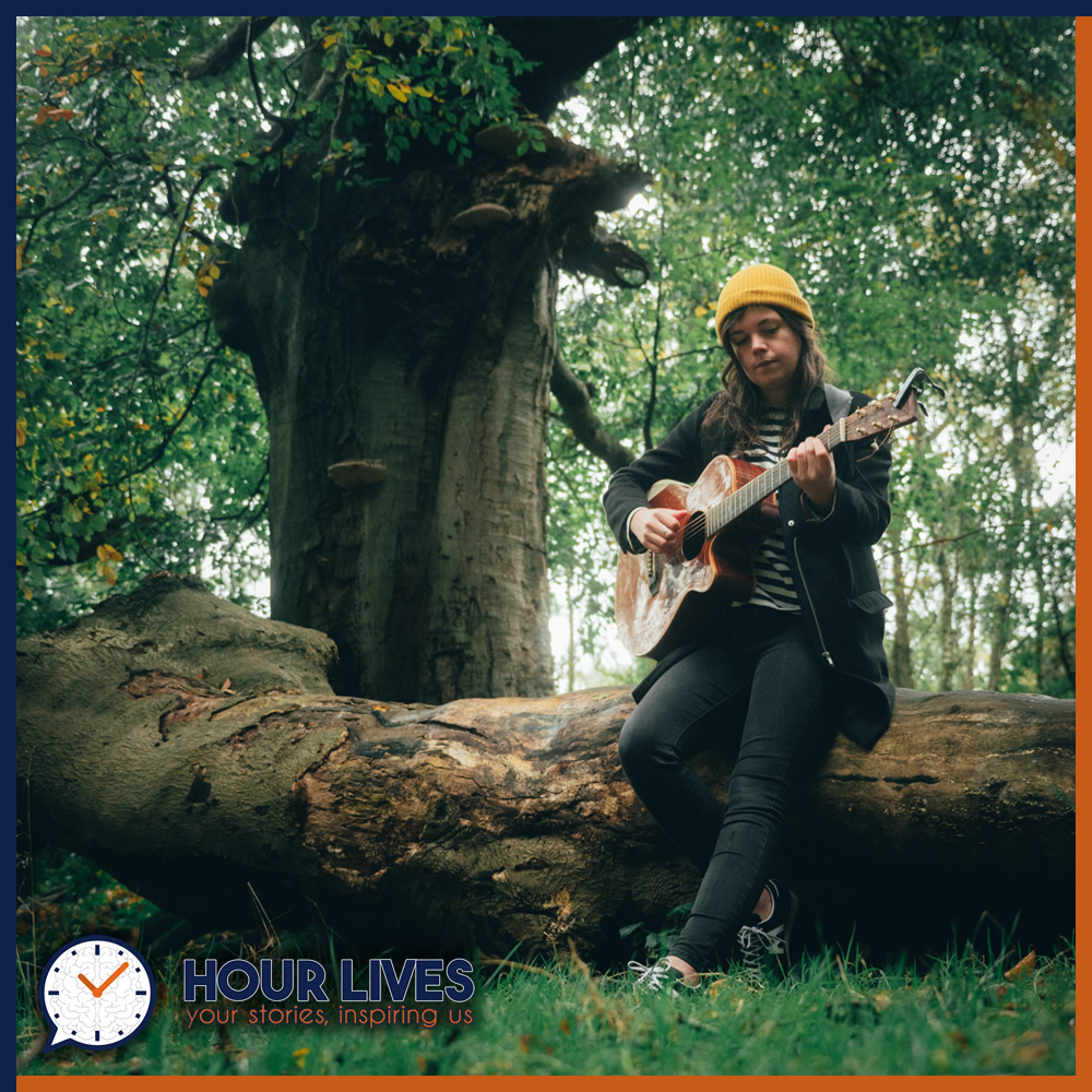 A photo of musician Jessie Reid, holding her guitar and perched on a fallen tree in a forest.